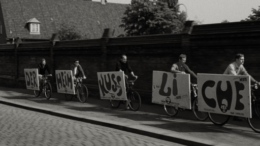Fünf Männer fahren auf Fahrrädern, an den Fahrrädern ist je ein beschriftetes Schild befestigt. Auf den Schildern steht DER, HEIM, KUSS, LI und CHE.