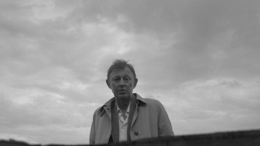 Ein Bild in schwarz-weiß. Ein älterer Mann steht vor einem bewölkten Himmel und schaut in die Richtung des Betrachters.