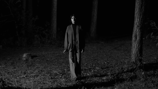 Das Bild ist in schwarz weiß. Eine junge Frau steht in einem dunklen Wald und schaut den Betrachter an.