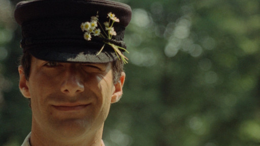 Ein Mann mit Mütze lächelt und zwinkert. An seiner Mütze sind Gänseblümchen befestigt.