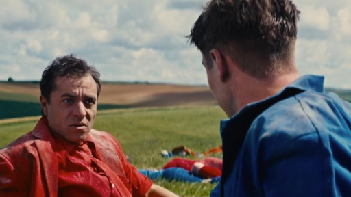 Zwei Männer auf einer Wiese, bewölkter Himmel. Ein Mann, blau bekleidet, ist von hinten zu sehen. Der andere Mann, in Rot gekleidet, schaut sein Gegenüber ernst an.